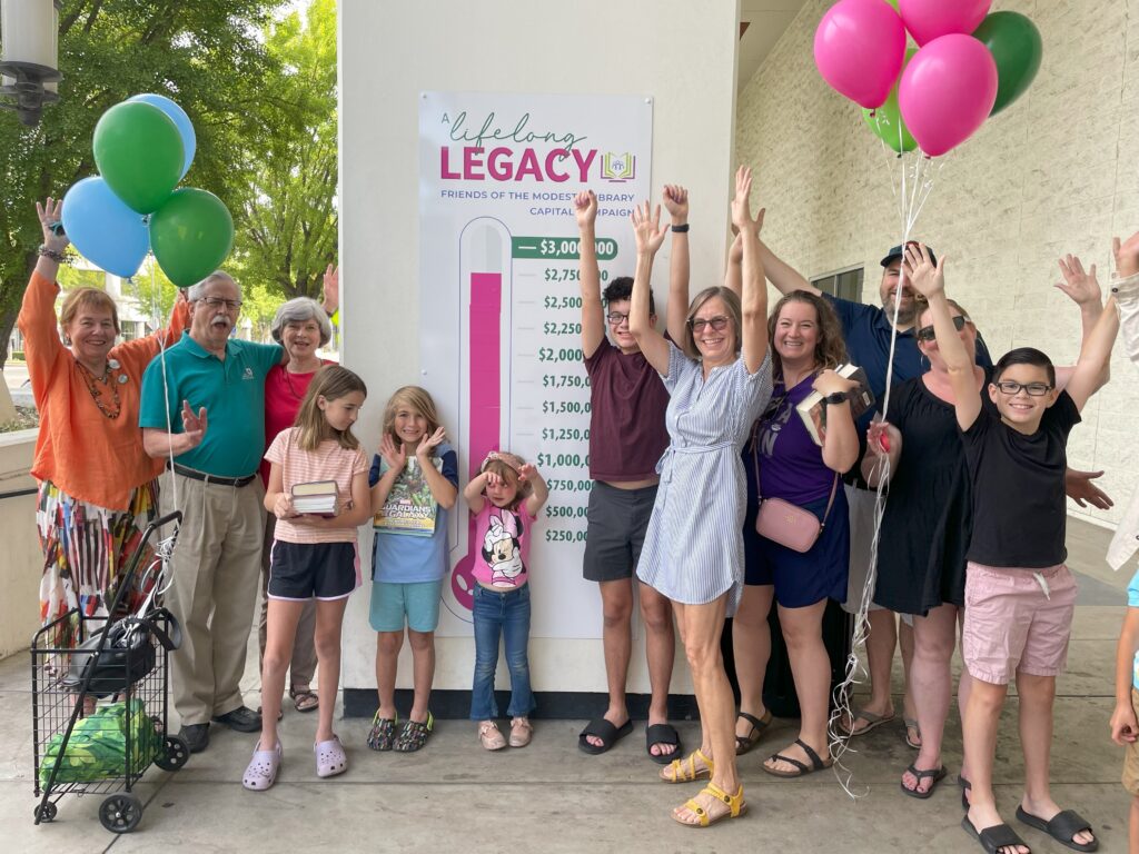 Children and adults hold balloons and raise their hands to celebrate while standing beside the campaign fundraising thermometer
