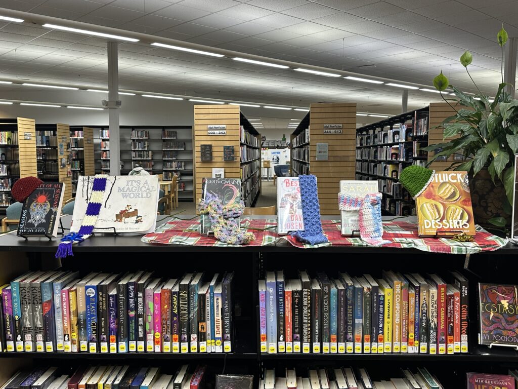 Books displayed on library shelves are adorned with handmade scarves and hats.