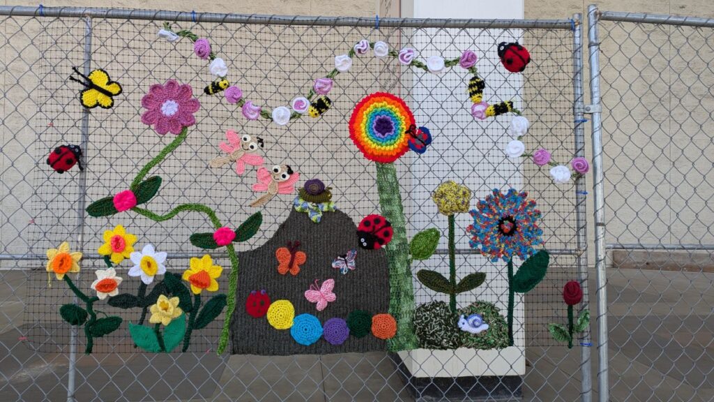 metal construction fence decorated with flowers, ladybugs butterflies and more all made of yarn