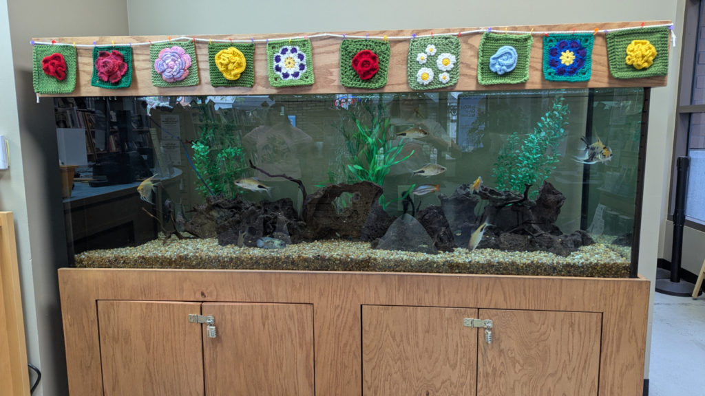 A colorful knitted and crocheted flower garland hangs at the top of the fish tank.